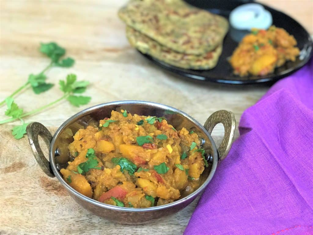 Instant Pot Pumpkin curry in a kadhai with cilantro for decoration. In the back, a plate with the pressure cooker pumpkin curry, parathas and onions. 