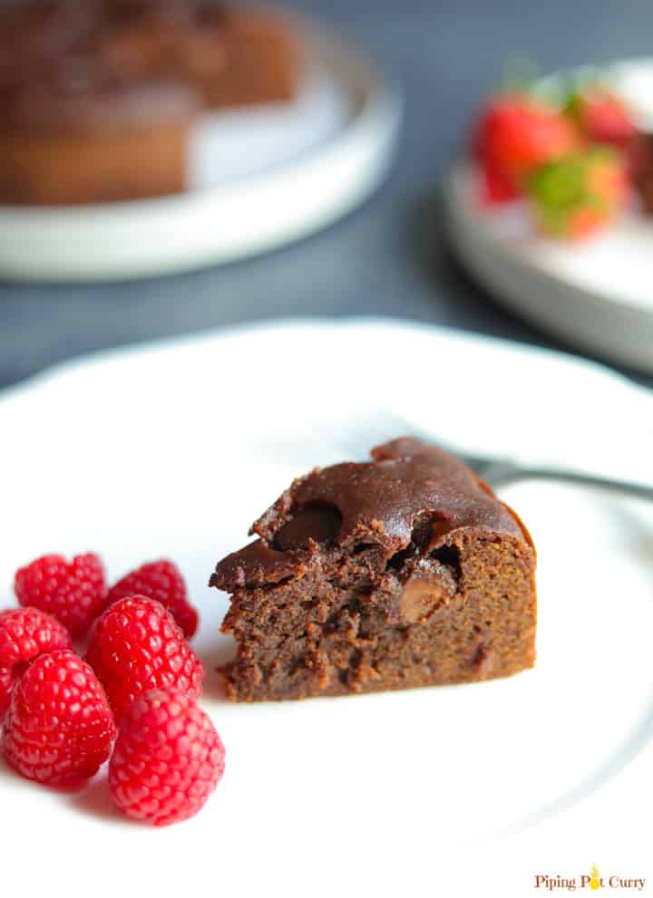 A piece of Instant Pot Brownie in a plate with raspberries, and the whole brownie cake in the back. 