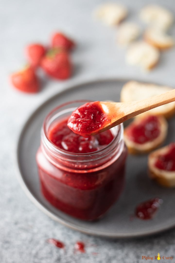 Spoonful of Strawberry Jam from a glass jar on a plate, along with jam spread on bread slices